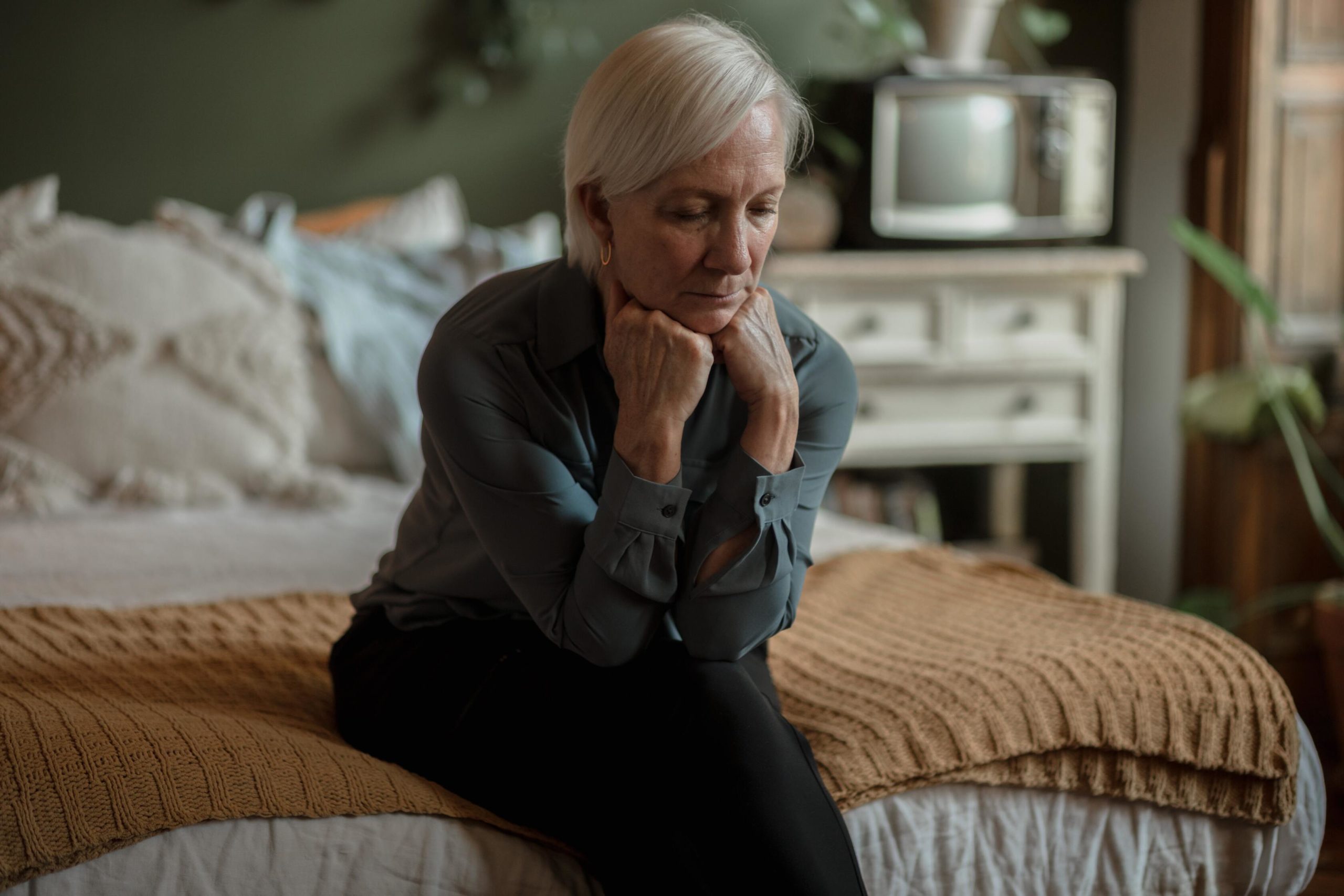 A senior woman sits on a bed alone.