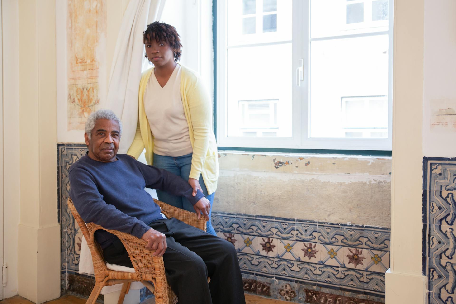 senior man sitting in chair with adult caretaker standing by his side